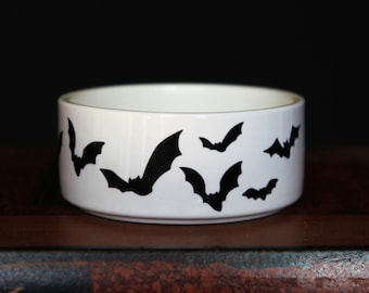 May include: A white ceramic bowl featuring a repeating pattern of black bat silhouettes around the exterior. The bowl has a smooth, cylindrical shape with a clean, white interior and a flat base, showcasing a high-contrast design.