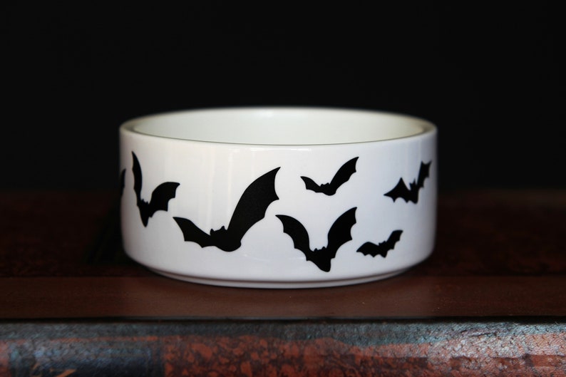 May include: A white ceramic bowl featuring a repeating pattern of black bat silhouettes around the exterior. The bowl has a smooth, cylindrical shape with a clean, white interior and a flat base, showcasing a high-contrast design.