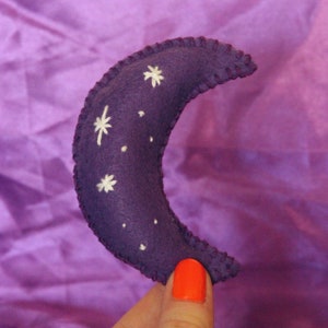 Pu&ograve; includere: Una mezzaluna di feltro viola con stelle bianche ricamate. La luna &egrave; tenuta in una mano con smalto per unghie arancione.