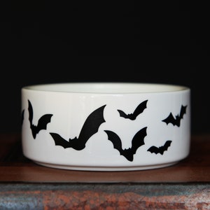 May include: A white ceramic bowl featuring a repeating pattern of black bat silhouettes around the exterior. The bowl has a cylindrical shape with a smooth, glossy finish and a flat base, providing a stark contrast between the dark bats and white.