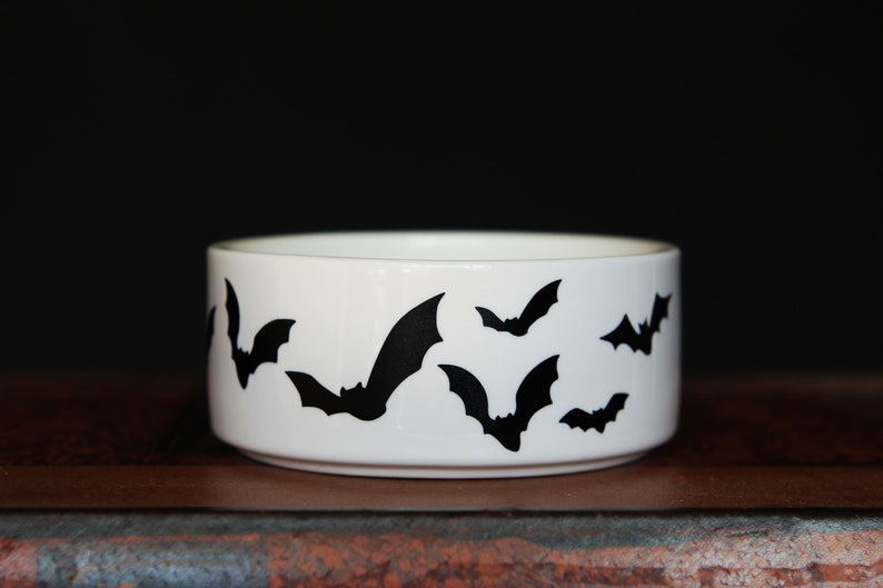 May include: A white ceramic bowl featuring a repeating pattern of black bat silhouettes around the exterior. The bowl has a cylindrical shape with a smooth, glossy finish and a flat base, providing a stark contrast between the dark bats and white.