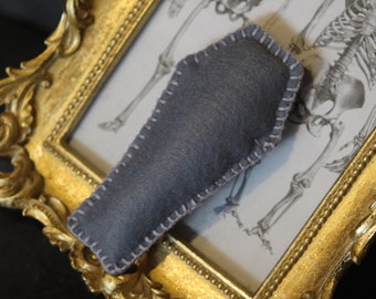 May include: A gray felt coffin-shaped pillow with white stitching, resting on a gold ornate picture frame with a black and white anatomical drawing of a skeleton inside.