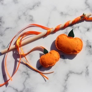 Op de afbeelding: Een houten toverstaf met oranje en gele lintjes en twee oranje viltpompoenen met groene stelen.