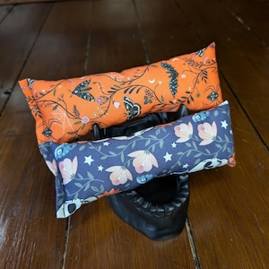 Op de afbeelding: Twee decoratieve kussens met bloemen- en insectenpatronen. Het bovenste kussen is oranje met zwarte insecten en witte bloemen. Het onderste kussen is blauw met witte sterren en roze bloemen.