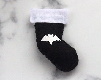 May include: A small black stocking with a white cuff and a white bat silhouette on the front. The stocking is likely for Halloween or a similar holiday. The background is a light, neutral color.