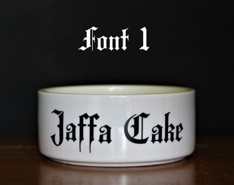 May include: A white ceramic bowl featuring the black text Jaffa Cake in a gothic-style font. Above the bowl, the text Font 1 is displayed in the same black gothic typeface. The bowl has a smooth, cylindrical shape with a clean, minimalist finish.