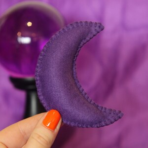Pu&ograve; includere: Cuscino a forma di mezzaluna in feltro viola con un bordo cucito.