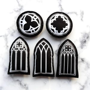 Peut inclure: Ensemble d'ornements en feutre noir et blanc de style gothique. L'ensemble comprend deux ornements circulaires et trois en forme de fenêtres cintrées. Les motifs blancs détaillés se détachent sur le fond noir, rappelant l'architecture des cathédrales.