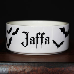 May include: A white ceramic bowl featuring the name Jaffa printed in a black gothic-style font. The exterior of the bowl is decorated with several black bat silhouettes scattered around the text, creating a high-contrast design on the white surface.