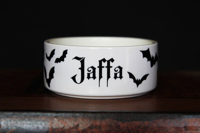 May include: A white ceramic bowl featuring the name Jaffa printed in a black gothic-style font. The exterior of the bowl is decorated with several black bat silhouettes scattered around the text, creating a high-contrast design on the white surface.