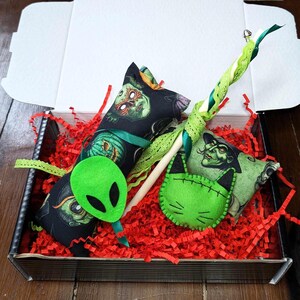 May include: A Halloween-themed cat toy set in a black box with red shredded paper. The set includes a green felt cat toy, a green felt alien toy, and a wand toy with a green ribbon and a wooden stick.
