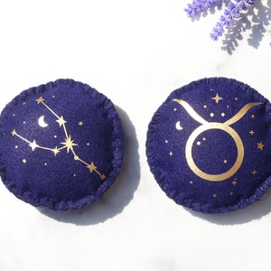 May include: Two round felt cushions featuring gold zodiac constellations. The first cushion is navy blue with a gold Taurus constellation. The second cushion is navy blue with a gold Gemini constellation.