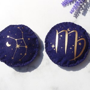 Op de afbeelding: Twee ronde vilten ornamenten met gouden borduurwerk. Een ornament toont het sterrenbeeld Maagd met een halve maan en sterren. Het andere ornament toont het astrologische symbool voor Maagd met sterren.