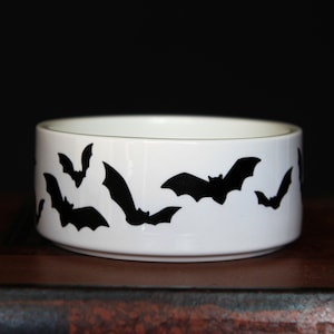 May include: A white ceramic bowl featuring a repeating pattern of black bat silhouettes around the exterior. The bowl has a cylindrical shape with a smooth finish and a clean, simple design, showcasing multiple bats in various flying positions.