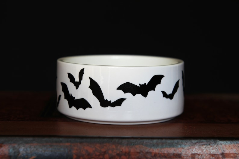 May include: A white ceramic bowl featuring a repeating pattern of black bat silhouettes around the exterior. The bowl has a cylindrical shape with a smooth finish and a clean, simple design, showcasing multiple bats in various flying positions.