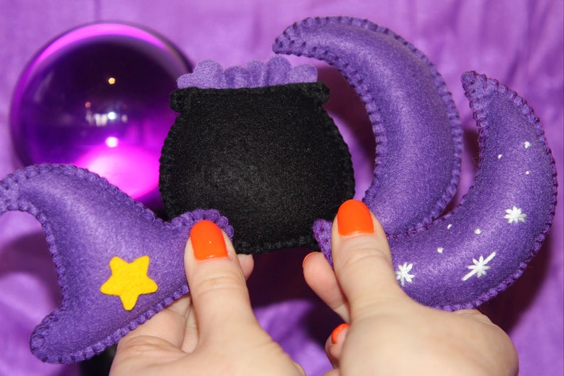 May include: A set of felt Halloween decorations featuring a black cauldron, a purple crescent moon with white stars, and a purple witch's hat with a yellow star.