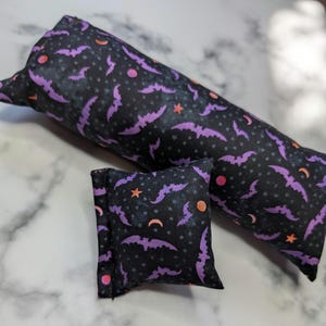 Puede incluir: Dos artículos de tela con temática de Halloween: una forma cilíndrica larga y una pequeña almohada cuadrada. Ambos son negros con un patrón repetido de murciélagos morados, lunas naranjas, estrellas y círculos rosas. Los artículos parecen hechos a mano.