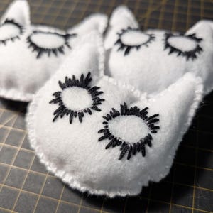 May include: White felt cat-shaped ornaments with black embroidered eyes and eyelashes. The ornaments are stitched with white thread around the edges. The cat-shaped ornaments are a craft item.