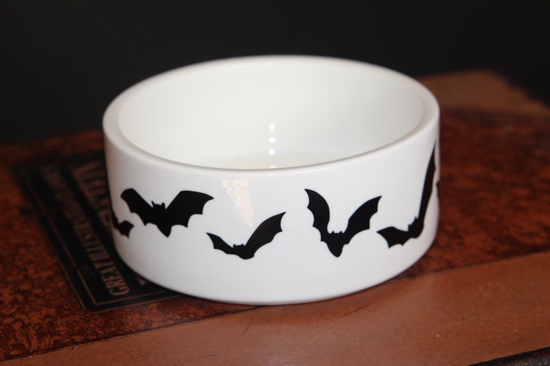 May include: A white ceramic bowl featuring a repeating pattern of black bat silhouettes around the exterior. The bowl has a smooth, glossy finish and a cylindrical shape with a wide, open top, providing a clear view of the interior basin.