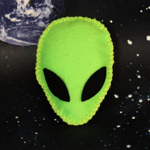 Könnte beinhalten: Grüner Filz-Alienkopf mit schwarzen Augen, mit gelbem Garn gestickt.