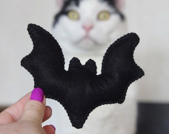 May include: A black felt bat toy with a stitched outline is held up in front of a blurred background. The bat has outstretched wings and a small head. The background features a white and black cat.