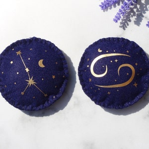 May include: Two round felt ornaments with gold zodiac signs on a white background. The first ornament features the constellation Cancer with a crescent moon. The second ornament features the Cancer zodiac symbol.