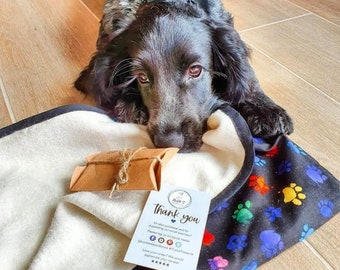 Könnte beinhalten: Ein schwarz-weißer Hund liegt auf einer schwarz-weißen Fleece-Decke mit bunten Pfotenabdrücken. Die Decke hat einen weißen Rand und eine Dankeskarte mit Social-Media-Informationen. Die Karte lautet: "Vielen Dank für Ihren Kauf, wir schätzen Ihre Unterstützung. Bitte taggen Sie uns in den sozialen Medien @duckandtreasure #duckandtreasure Gefällt Ihnen Ihre Bestellung? Wir würden uns über eine 5-Sterne-Bewertung freuen."