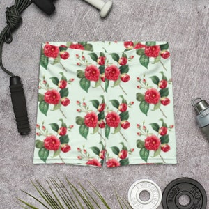 Op de afbeelding: Een paar sport shorts met een bloemenprint op een lichtgroene achtergrond en rode bloemen. De shorts liggen op een grijs oppervlak met een springtouw, gewichten en een waterfles.