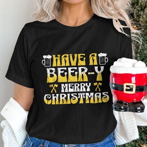 以下が含まれることがあります： 「Have a Beer-y Merry Christmas」と黄色いグラフィックが書かれた黒いTシャツ。グラフィックには、ビールジョッキ2つと交差した斧2つが含まれています。
