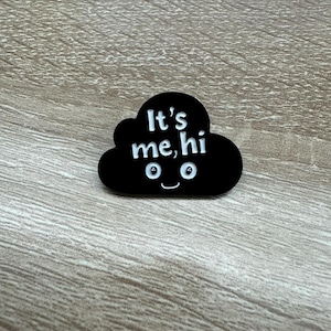 May include: Black enamel pin shaped like a cloud with white text that says "It's me, hi" and two white eyes with a white smile.