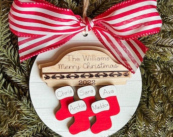 May include: A white wooden Christmas ornament with a red and white striped ribbon. The ornament features a car silhouette with the text "The Williams Merry Christmas 2022" and five stockings with the names "Chris", "Sara", "Ava", "Archie", and "Maddox".