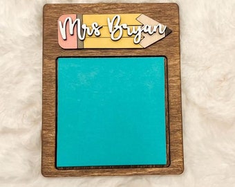 May include: A wooden sign with a cutout of a pencil with the text "Mrs. Bryan" on it. The sign has a turquoise blue dry erase board in the center.