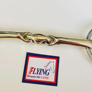 May include: A horse bit featuring stainless steel rings and a gold-toned, twisted central section. A small tag with the word "FLYING" and a horse head logo is also present.