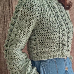 Pode incluir: Um suéter de crochê verde-sálvia com um padrão de tricô trançado e mangas compridas. O suéter é cropped e tem um caimento relaxado.