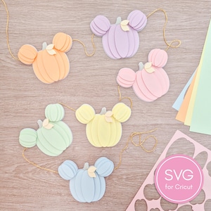 May include: A garland featuring six layered paper pumpkins in pastel shades of orange, lavender, pink, mint green, yellow, and light blue, each with mouse-ear shapes and a small cream leaf, strung on gold cord with a pink SVG for Cricut label.