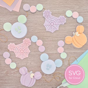 Puede incluir: Una guirnalda decorativa con recortes de papel en capas con formas de fantasmas, telarañas y calabazas en tonos pastel lavanda, rosa, verde menta y naranja, unidos por un cordel dorado. Una etiqueta circular rosa muestra el texto SVG for Cricut.