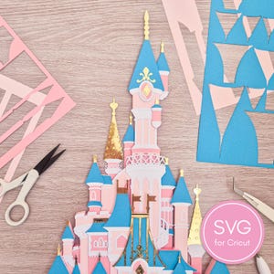 May include: A layered paper castle in pink, blue, and gold, with intricate cut details. The castle is surrounded by cut paper pieces and tools, including scissors and tweezers. A pink circle with the text "SVG for Cricut" is in the lower right.