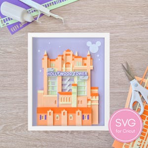 May include: A framed paper art piece depicting the Hollywood Tower Hotel in shades of orange, peach, and purple. The artwork includes the hotel's name and a Mickey Mouse head silhouette. Additional crafting supplies are visible.