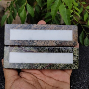 May include: Two rectangular stone blocks with a white rectangular inlay. The stone has a mottled pattern of green, brown, and grey. The blocks are held in a hand, with green foliage in the background.