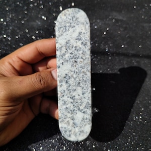 Peut inclure: Un objet en pierre rectangulaire, moucheté de gris et de blanc, avec des extrémités arrondies. La pierre a une surface polie et est tenue dans une main sur un fond sombre et scintillant. L'objet est long et étroit.