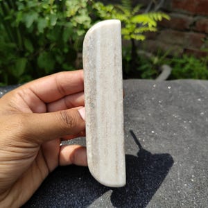 May include: A light-colored, rectangular stone massage tool with rounded edges. The stone has a natural, textured appearance with streaks of gray and beige. The tool is held in a hand against a blurred background of greenery and a brick wall.
