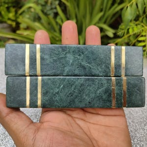 May include: Two rectangular, dark green marble bookends with gold-colored vertical stripes. The bookends are held in a hand, with a natural background of green foliage. The marble has a natural veining pattern.