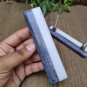 May include: A sharpening stone with a white, gray, and black color scheme. The stone is rectangular and has a metal handle. The stone is designed for sharpening knives and other tools. The stone is held in a hand.