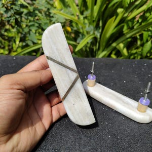 May include: Two elongated, semi-cylindrical cabinet pulls crafted from polished white marble with subtle grey veining. Each handle features a distinct diagonal inlay of dark grey stone. The pulls include metal mounting bolts and purple spacers.