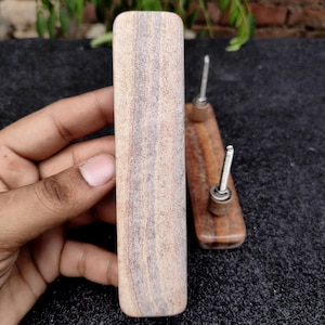 May include: A light-coloured wooden drawer pull held in a hand, with a rectangular shape and visible wood grain. Two smaller, similar drawer pulls with metal screws are also visible. The pulls are likely for furniture or cabinetry.