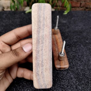 Op de afbeelding: Close-up van twee houten kastknoppen met metalen schroeven. Eén knop wordt in een hand gehouden, die de natuurlijke houtnerf laat zien. De andere knop ligt op een donkere ondergrond. De knoppen zijn rechthoekig en cilindrisch.