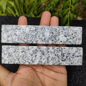 May include: Two rectangular granite stones with a speckled pattern of white, gray, and black. The stones are held in a hand, showcasing their polished surface and natural color variations. These stones could be used for home decor or craft projects.