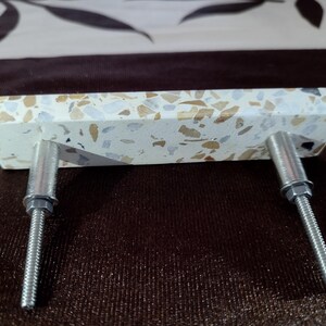 May include: A rectangular cabinet pull with a speckled, off-white surface. The pull features flecks of brown, beige, and grey. Two silver metal bolts extend from the back for mounting. This hardware is suitable for drawers and cabinets.