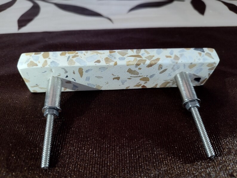 May include: A rectangular cabinet pull with a speckled, off-white surface. The pull features flecks of brown, beige, and grey. Two silver metal bolts extend from the back for mounting. This hardware is suitable for drawers and cabinets.