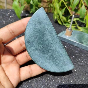 May include: A semi-circular, teal-colored stone with a textured surface. The stone is held in a hand, with a matching stone base with metal posts in the background. The stone appears to be a decorative item or a component of a larger piece.